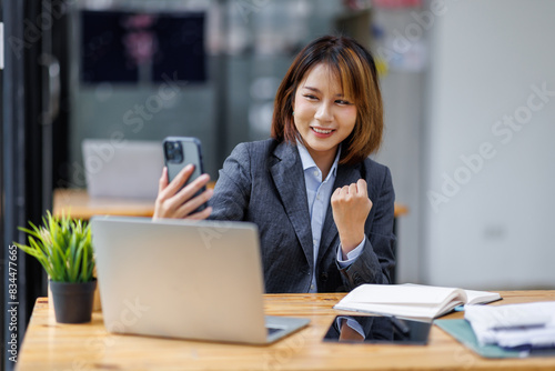 Young busy smart serious Asian Japanese business woman executive holding cellphone device using mobile cell phone looking at smartphone working in corporate office with laptop computer technology.