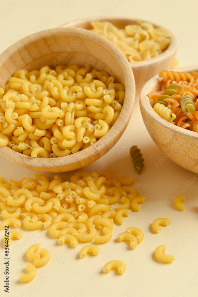 Wooden bowls with raw elbow and fusilli pasta on beige background, closeup Wooden bowls with raw elbow and fusilli pasta on beige background, closeup