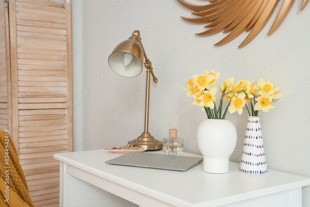 © Pixel-Shot - Vase with narcissus flowers, lamp and laptop on table in stylish room