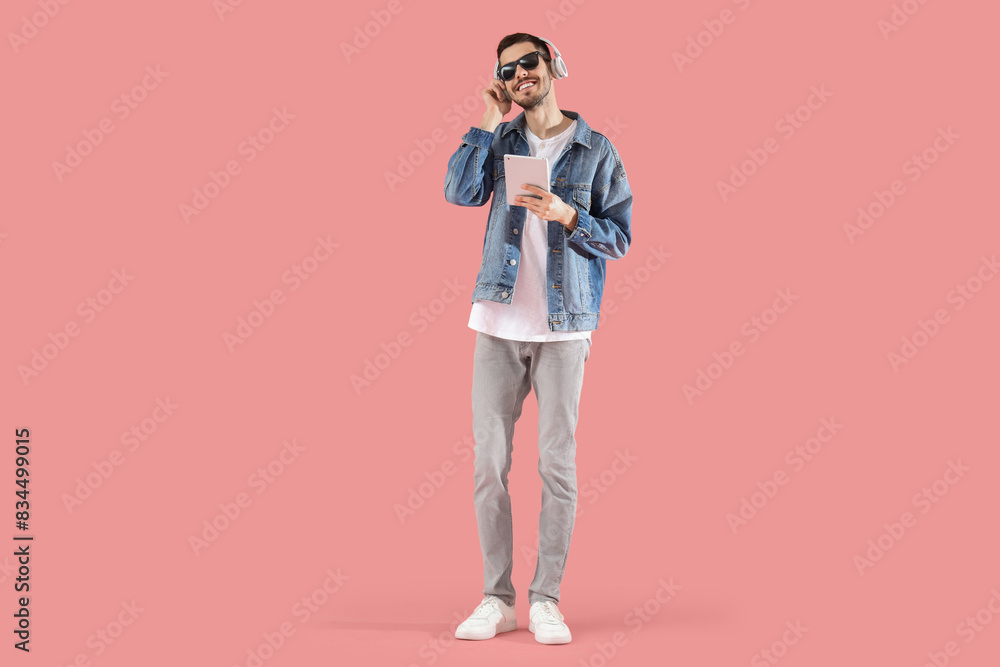 Fototapeta premium Young bearded man in headphones with tablet computer listening to music on pink background