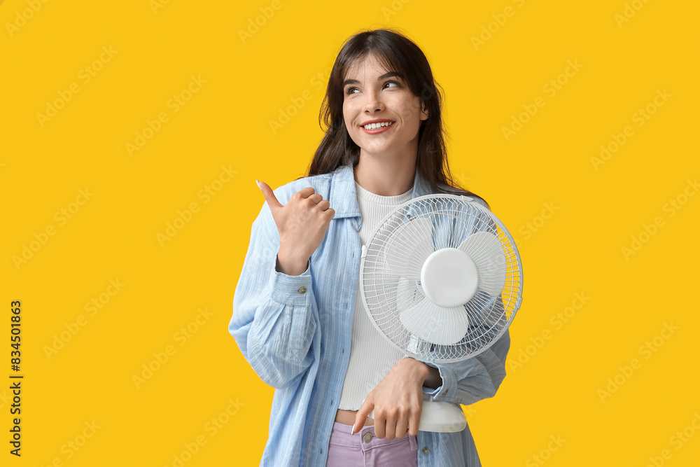 Young woman with electric fan pointing at something on yellow background Young woman with electric fan pointing at something on yellow background