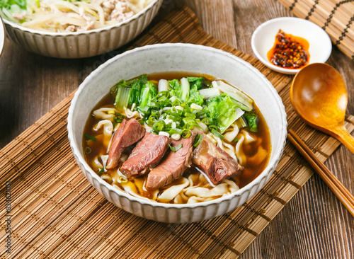 Classic Beef Tendon Noodles with chilli sauce, spring onion, wooden spoon, and chopsticks served in dish isolated on mat side view of taiwanese food