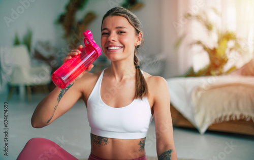 Photo of a satisfied slim woman in sportswear sitting on the floor after exercises or gym looking on camera with smile and drinking water from a pink sports bottle