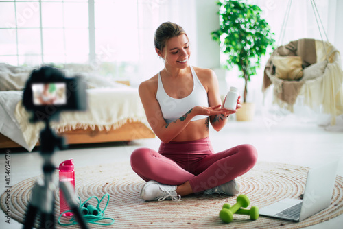 Cute slim sporty girl blogger in sportfit sitting on the floor with laptop and dumbbells and showing a white jar of sports nutrition protein supplements to the camera at home photo