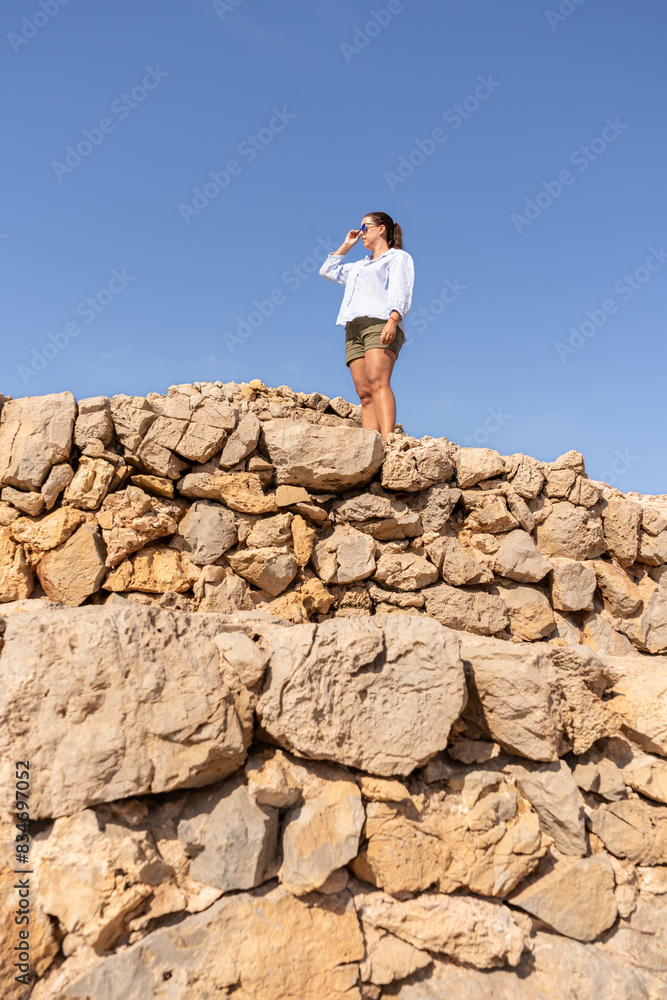 A woman in her 30 in sunglasses looking forward wearing casual outfit stands confidently on ancient stone walls against a clear blue sky A woman in her 30 in sunglasses looking forward wearing casual outfit stands confidently on ancient stone walls against a clear blue sky