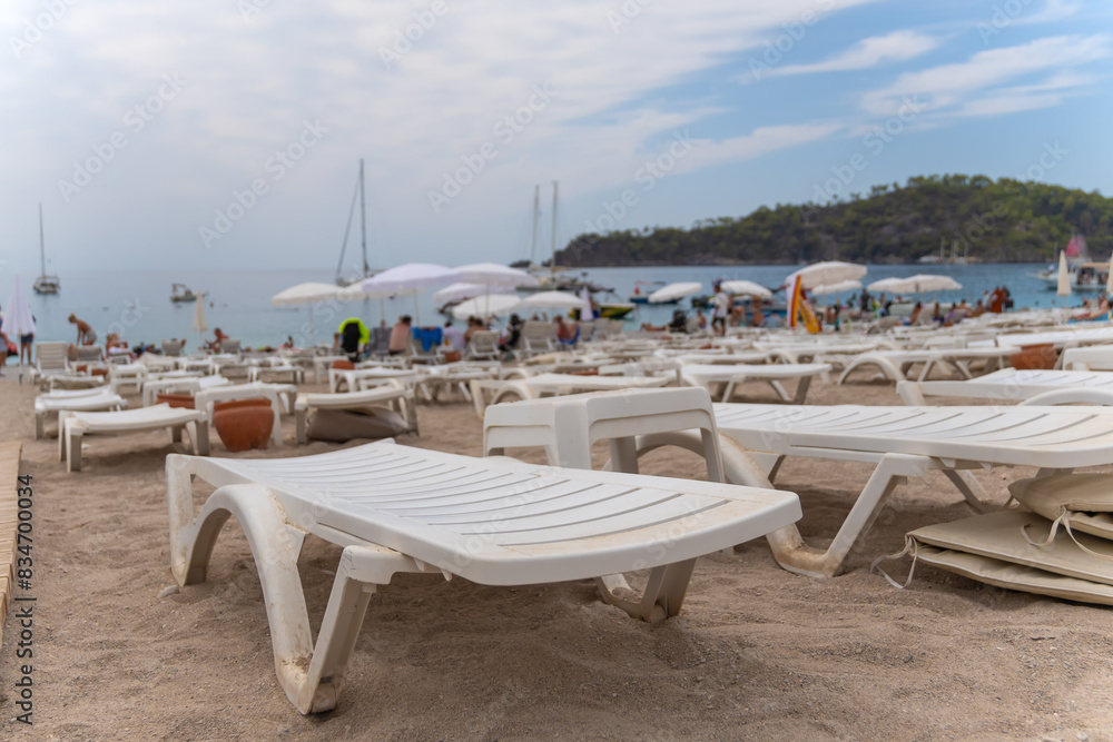 Close-up of white plastic sunbeds on the beach against the background of the blurred sea, mountains, yachts and boats. Close-up of white plastic sunbeds on the beach against the background of the blurred sea, mountains, yachts and boats.