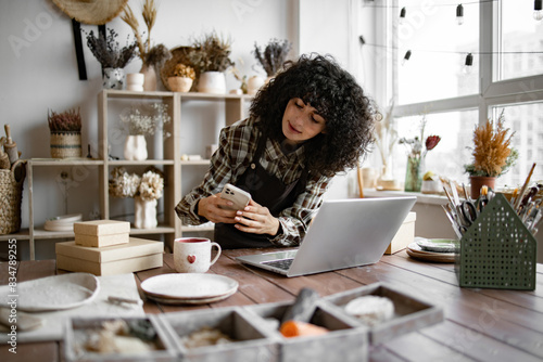 Charismatic Caucasian woman seller in souvenir shop takes pictures of goods at phone for posting on social networks. Successful girl store employee with ceramic tableware shoots photos on smartphone.