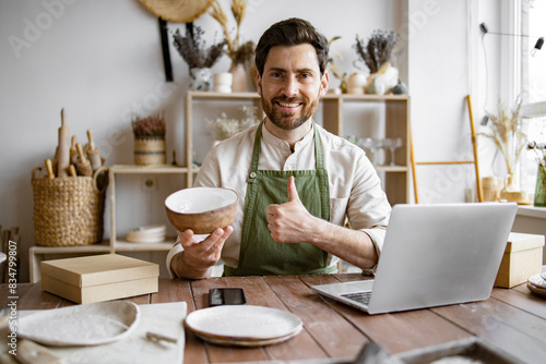 Happy worker in apron holds unique bowl created in personal style against background of shelves with dishes. Adult bearded male potter engaged in e-commerce selling handmade clay products on Internet.
