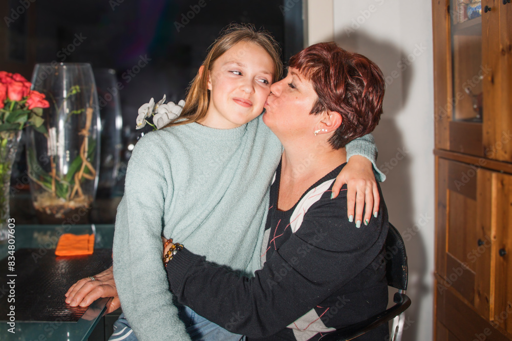 Obraz premium Positive emotions at a family holiday at home. A woman with a teenage girl on her lap gives a kiss