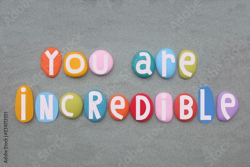 You are incredible, creative and motivational text composed with hand painted multi colored stone letters over green sand