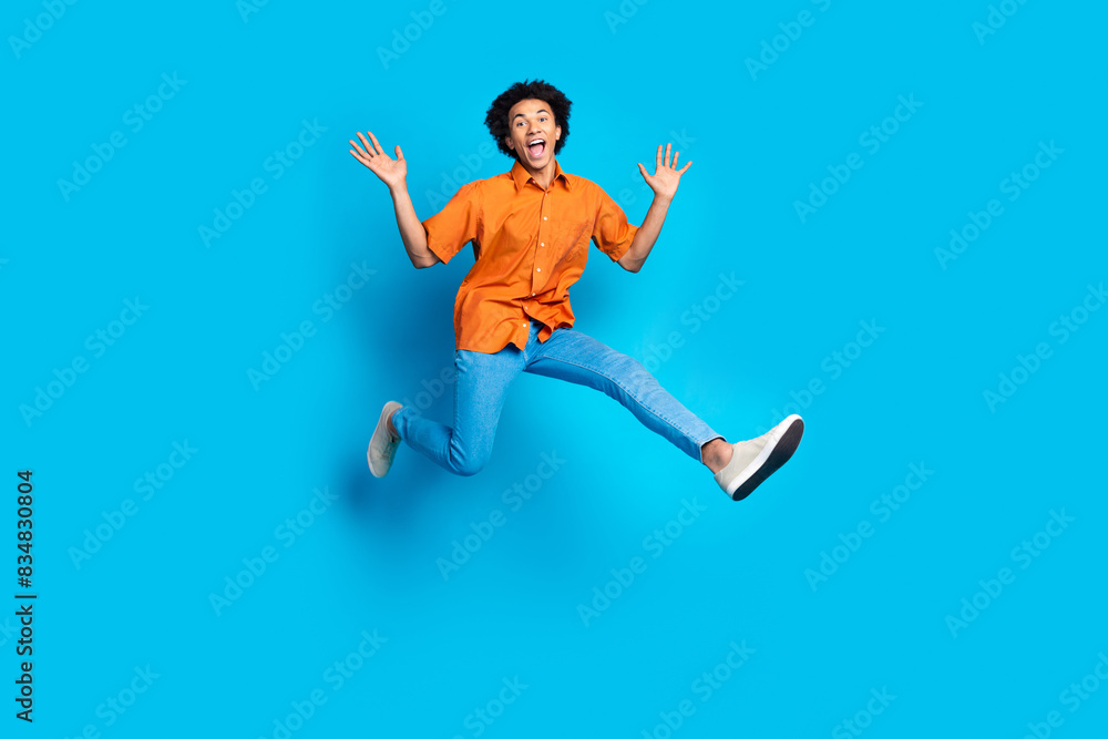 Full length photo of handsome good mood guy wear orange shirt jumping high emtpy space isolated blue color background Full length photo of handsome good mood guy wear orange shirt jumping high emtpy space isolated blue color background