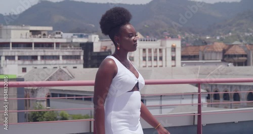 In a white dress, a young black girl stands on a rooftop with a view of the city. photo