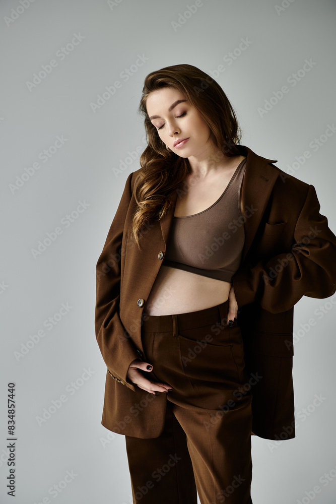 A young, pregnant woman in a stylish brown suit with a blazer confidently posing on a grey background. A young, pregnant woman in a stylish brown suit with a blazer confidently posing on a grey background.