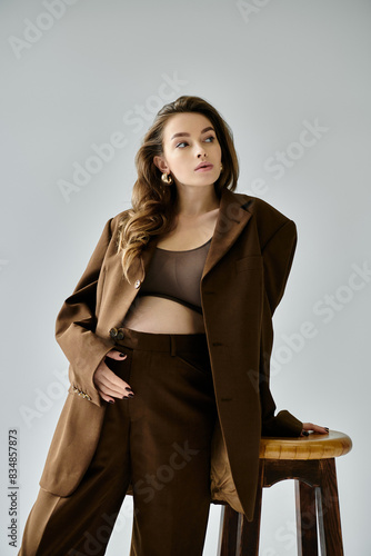 A young pregnant woman in a brown suit with a blazer sitting elegantly on top of a wooden stool against a grey background.