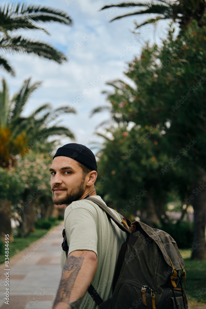 Follow me travel concept. A young man with a retro-style green backpack stands on an alley with palm trees in a subtropical climate. Montenegro, Bar town resort. Happy face and hand with tattoo. Follow me travel concept. A young man with a retro-style green backpack stands on an alley with palm trees in a subtropical climate. Montenegro, Bar town resort. Happy face and hand with tattoo.