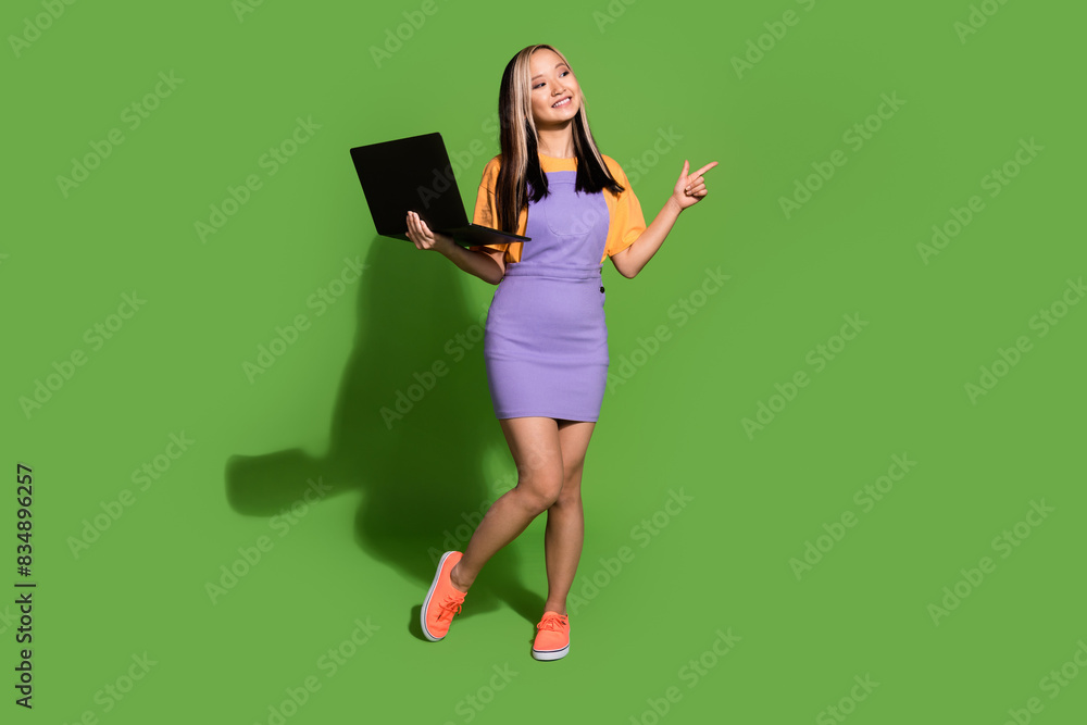 Full size photo of pretty young girl hold laptop point look empty space wear trendy violet yellow outfit isolated on green color background Full size photo of pretty young girl hold laptop point look empty space wear trendy violet yellow outfit isolated on green color background