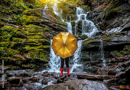Woman in red rubber boots behind yellow umbrella on mountains waterfall