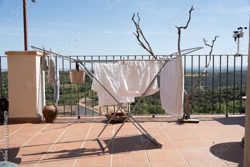 Photograph of washing clothes, sheets and towels on a clothesline on a terrace, in the background views of a village surrounded by fields and woods.