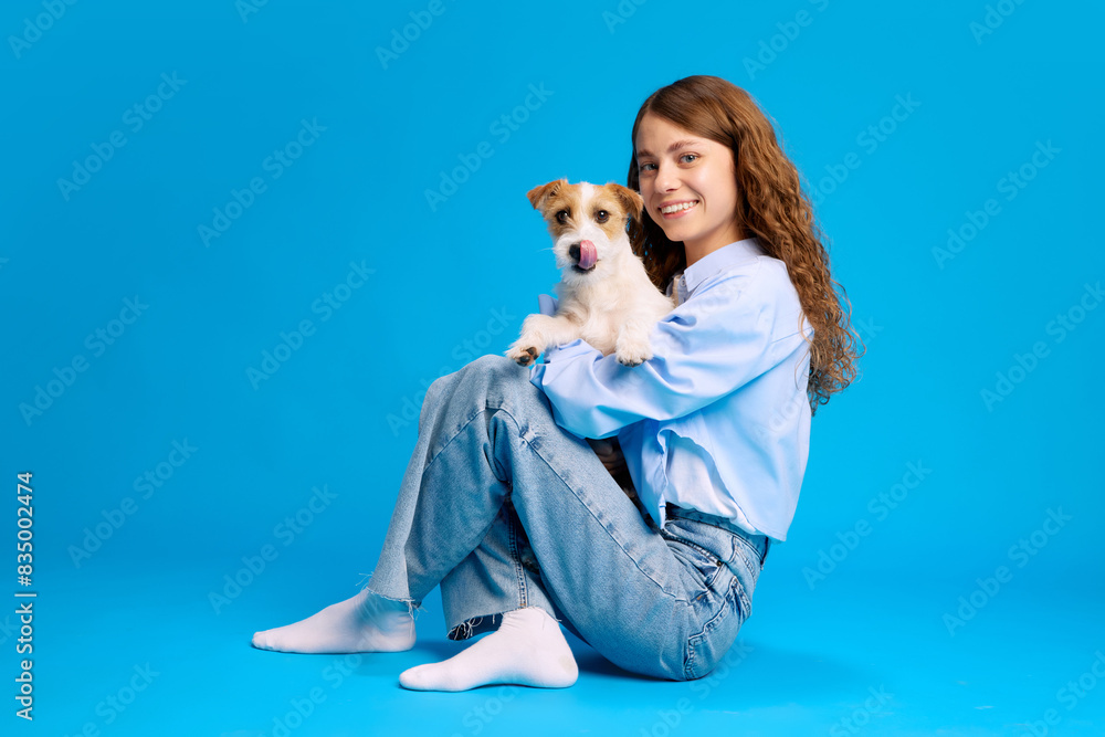 Portrait of young woman sits with cute purebred jack Russel puppy against vibrant blue background, enjoying moment. Concept of animals and their owners, friendship, pets care. Ad Portrait of young woman sits with cute purebred jack Russel puppy against vibrant blue background, enjoying moment. Concept of animals and their owners, friendship, pets care. Ad