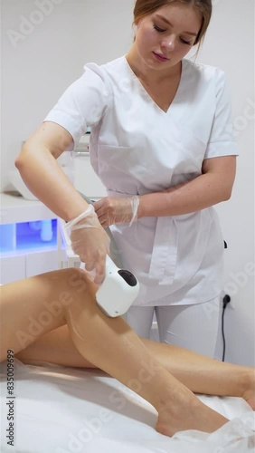 Female cosmetologist performing laser hair removal depilation on slender legs of stunning elegant lady in black bodysuit lying on medical couch in beauty salon. Vertical orientation. Cosmetology