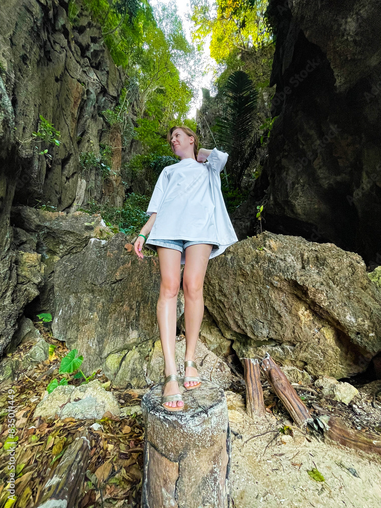 A woman in an oversized white t-shirt and shorts stands on a tree stump in a lush forest, surrounded by tall cliffs and greenery, looking serene and contemplative. A woman in an oversized white t-shirt and shorts stands on a tree stump in a lush forest, surrounded by tall cliffs and greenery, looking serene and contemplative.