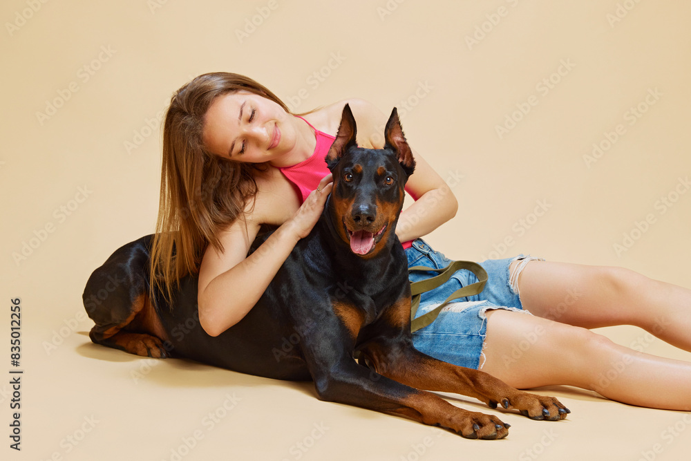 Young woman in pink top and jean shorts lies on floor cuddling with her large Doberman against beige studio background. Concept of animals and their owners, friendship, pets care. Ad Young woman in pink top and jean shorts lies on floor cuddling with her large Doberman against beige studio background. Concept of animals and their owners, friendship, pets care. Ad