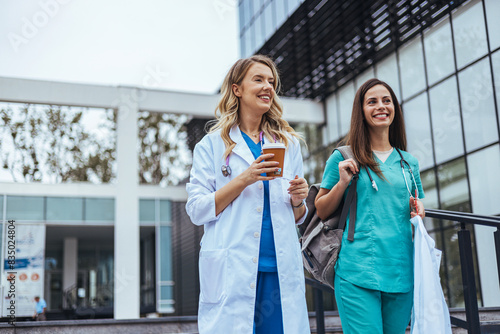 Bild auf Leinwand Two cheerful Caucasian women in healthcare attire, a doctor and nurse, walk and converse outside a medical facility, exuding relaxation after a workday
