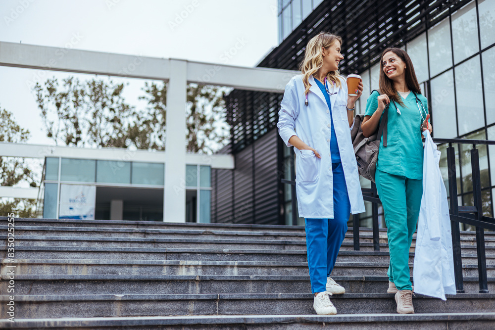Two cheerful Caucasian women in healthcare attire enjoy a light conversation while descending stairs outside a medical facility, a coffee cup in one's hand signals a relaxed end to their workday.