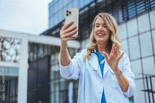 A cheerful Caucasian female healthcare worker in a white lab coat engages with her smartphone outside a medical facility, gesturing a friendly wave during a video call.