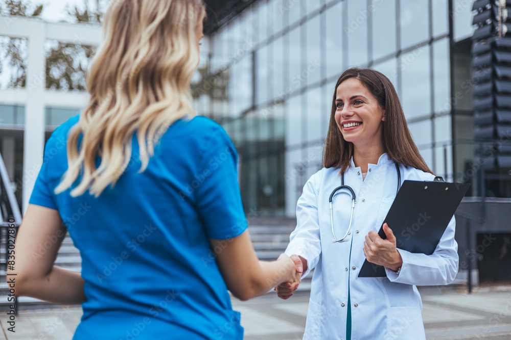 Two Caucasian female healthcare workers collaborate outside a medical facility; one in a lab coat holds a clipboard as they engage in a handshake. Two Caucasian female healthcare workers collaborate outside a medical facility; one in a lab coat holds a clipboard as they engage in a handshake.