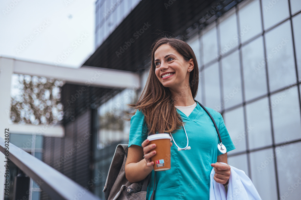 © Dragana Gordic - A cheerful Caucasian female nurse, clad in scrubs with a stethoscope, relishes a break while holding a coffee cup, standing before a medical facility.