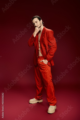 A handsome man in a red suit poses confidently without a shirt in a studio setting.