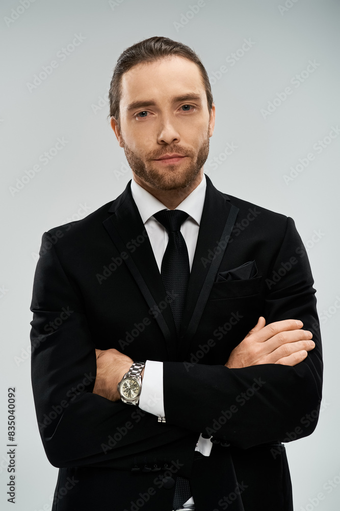 A bearded businessman in a sleek suit and tie confidently crosses his arms against a grey studio backdrop.