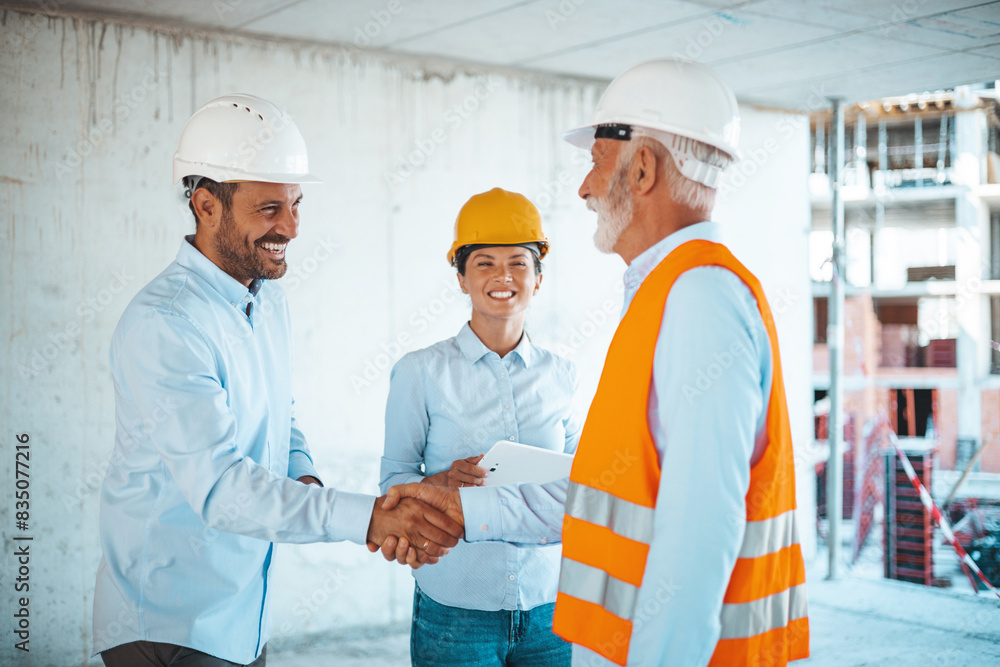 Two male construction site managers, representing diverse ethnicities, shake hands on a building site, with a smiling female colleague observing, all in safety gear.