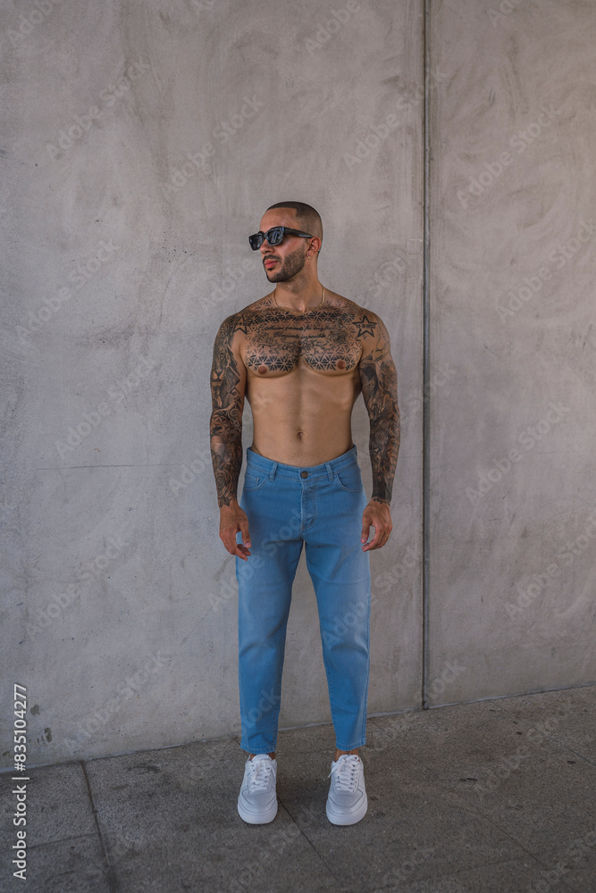 Chico joven musculoso y tatuado posando con vaqueros frente a pared Chico joven musculoso y tatuado posando con vaqueros frente a pared