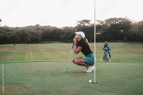 Obraz na plátně A young woman a beginner golfer, is squatting on the golf course and holding a club in her hands