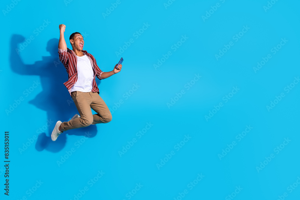 Fototapeta premium Full length photo of ecstatic guy wear stylish t-shirt hold smartphone jump win lottery look empty space isolated on blue color background