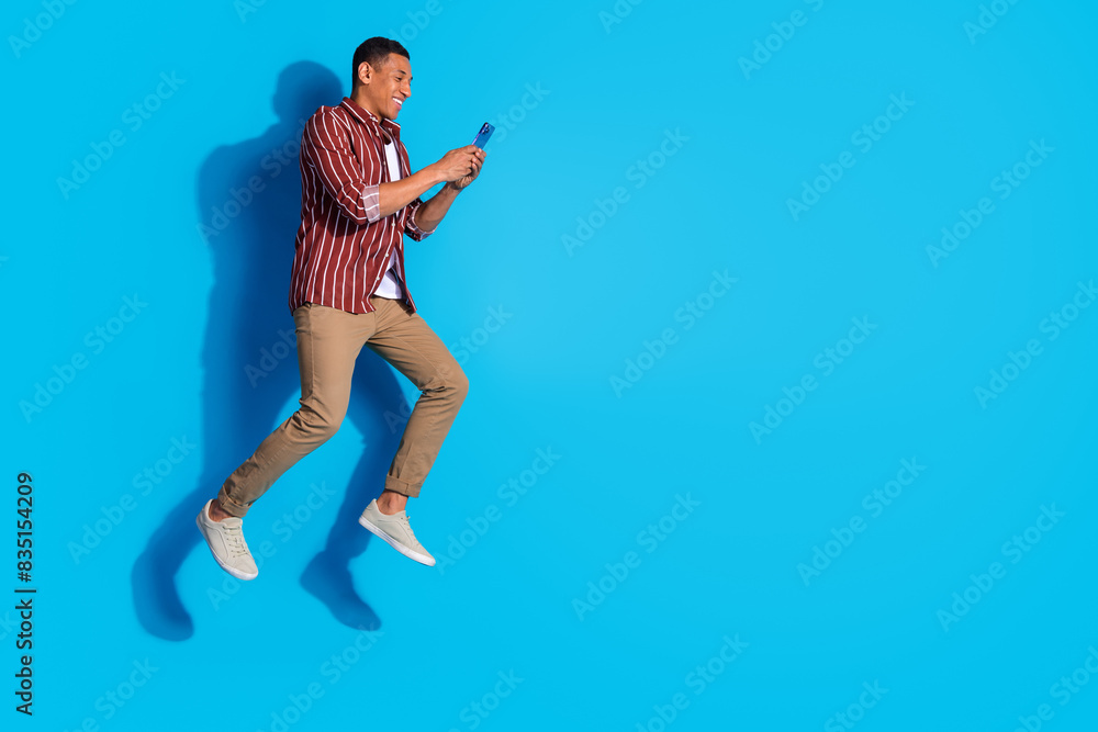 Full length photo of positive guy wear stylish t-shirt pants look at smartphone fly to empty space isolated on blue color background