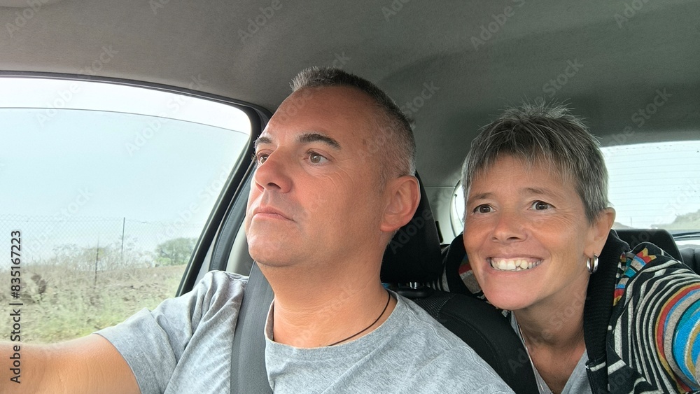Young adult couple sitting in car, he in the driver's seat looking straight ahead, she in the back seat, leaning between the two front seats, looking out the front window. Traveling with friends. Young adult couple sitting in car, he in the driver's seat looking straight ahead, she in the back seat, leaning between the two front seats, looking out the front window. Traveling with friends.