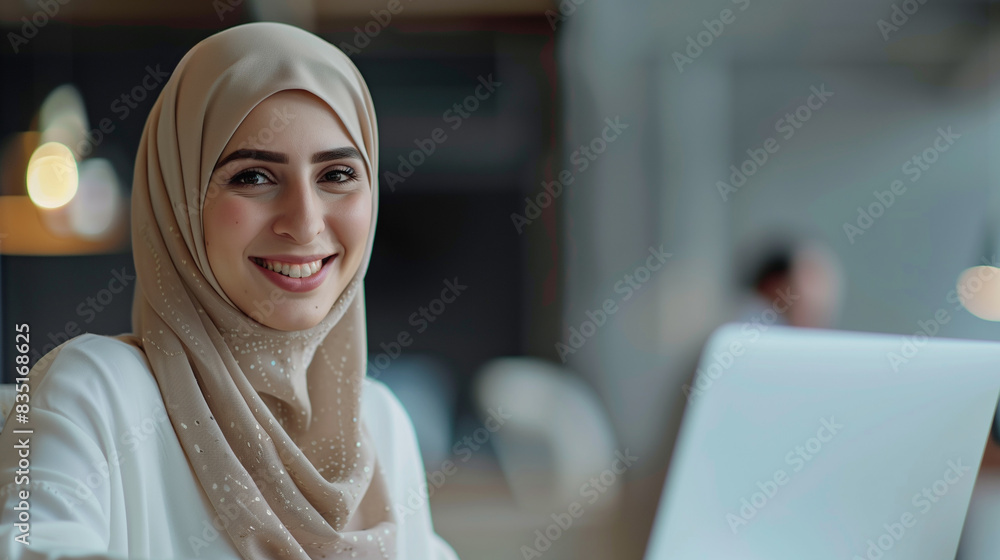 Muslim woman in a hijab working on a laptop at a modern office, smiling, blurred gray background Muslim woman in a hijab working on a laptop at a modern office, smiling, blurred gray background