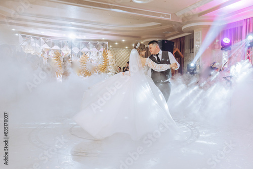 A bride and groom are dancing in a ballroom with a fog machine. The bride is wearing a white dress and the groom is wearing a suit. The fog machine creates a misty atmosphere, adding to the romantic