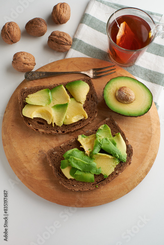 A close-up of a breakfast meal consisting of two slices of avocado toast, a cup of tea, a red apple, and walnuts on a wooden cutting board.