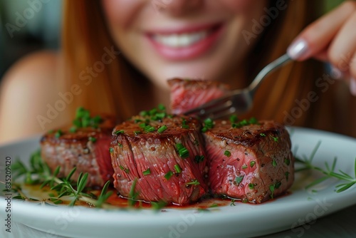 Carnivore diet concept. Happy young woman with smile on her face eats rare or medium-rare beef steak from a white plate at the table using fork and knife closeup portrait.
