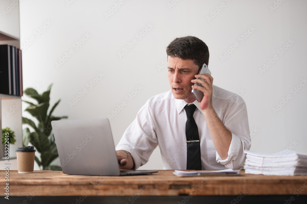 Businessman on phone call while working on laptop with financial reports, concept of multitasking and communication Businessman on phone call while working on laptop with financial reports, concept of multitasking and communication