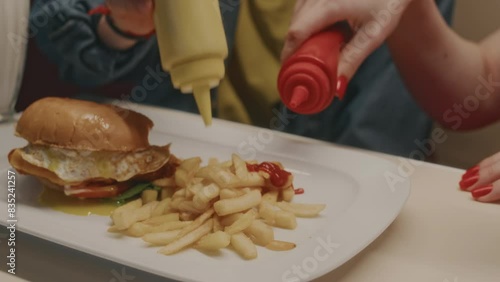 Closeup of anonymous customers pouring mustard and ketchup over fries while eating in cafe, having supper with fries and burger