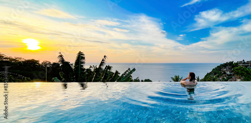 A woman enjoys a serene moment in an infinity pool overlooking the ocean at sunset. The lush tropical scenery and peaceful ambiance create a perfect setting for themes of relaxation, luxury, and