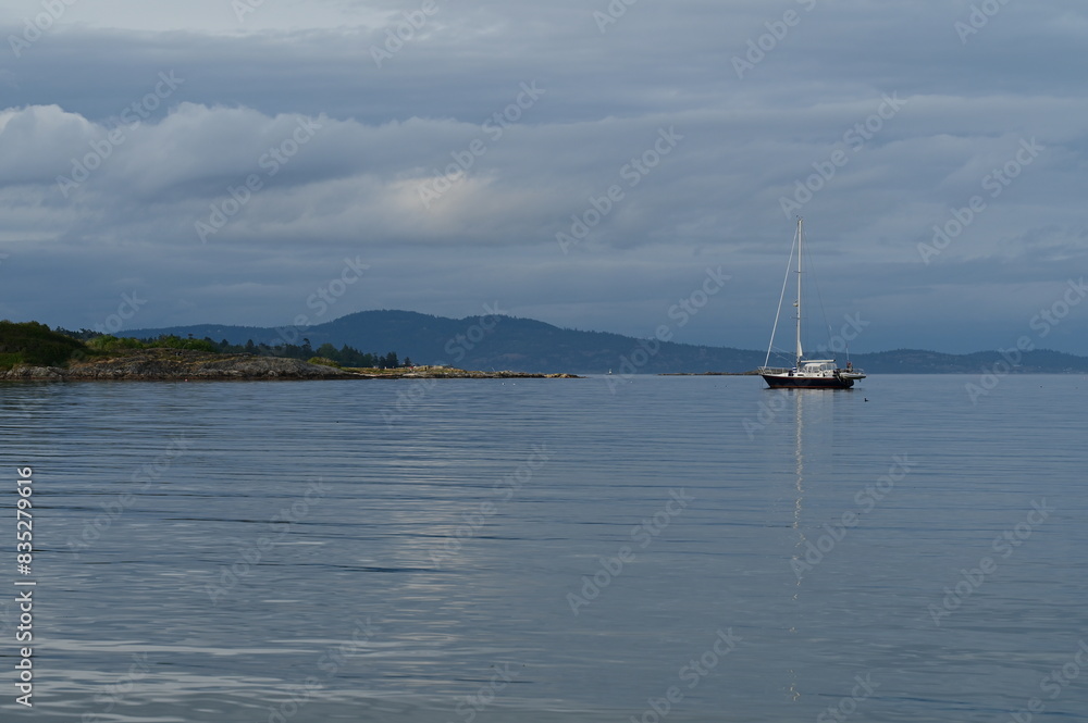 a sail boat on water under clouds and trees near an island a sail boat on water under clouds and trees near an island
