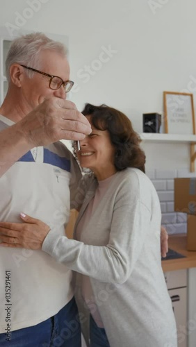 Happy senior man and woman show key to long-awaited flat at camera coming in kitchen. Elderly couple feels happy standing near paper boxes from POV to third party Vertical Shot. photo