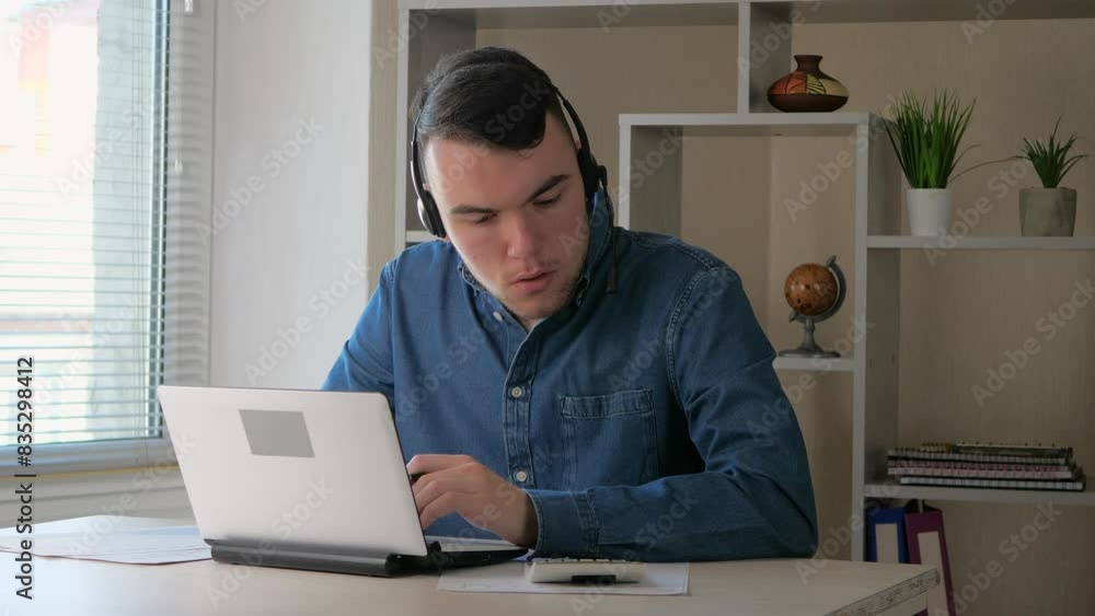 A young businessman, wearing headphones, is working on compiling an accounting report, communicating with colleagues and reconciling data using a calculator and entering them into the computer. A young businessman, wearing headphones, is working on compiling an accounting report, communicating with colleagues and reconciling data using a calculator and entering them into the computer.