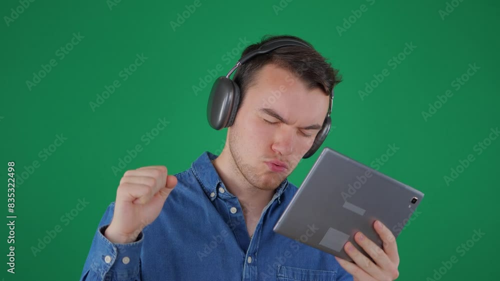 A young handsome man in headphones and a blue shirt listens to music with a tablet in his hands, swaying to the beat and gesturing with his hands. Green background. Slow motion. A young handsome man in headphones and a blue shirt listens to music with a tablet in his hands, swaying to the beat and gesturing with his hands. Green background. Slow motion.